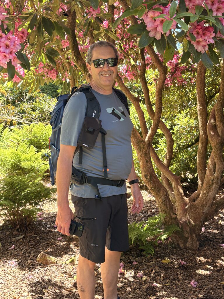 Kurt Locher steht mit grossem Wanderrucksack und Sonnenbrille vor einem blühenden Rhododendron mit rosa Blüten