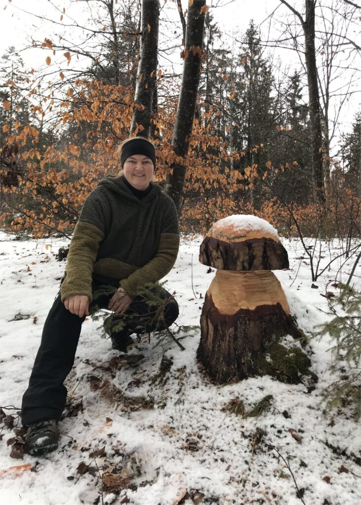 Sarah Hess sitzt in olivgrüner Fleecejacke und dunkler Mütze im verschneiten Wald neben einer aus einem Baumstrunk geschnitzten Pilzfigur