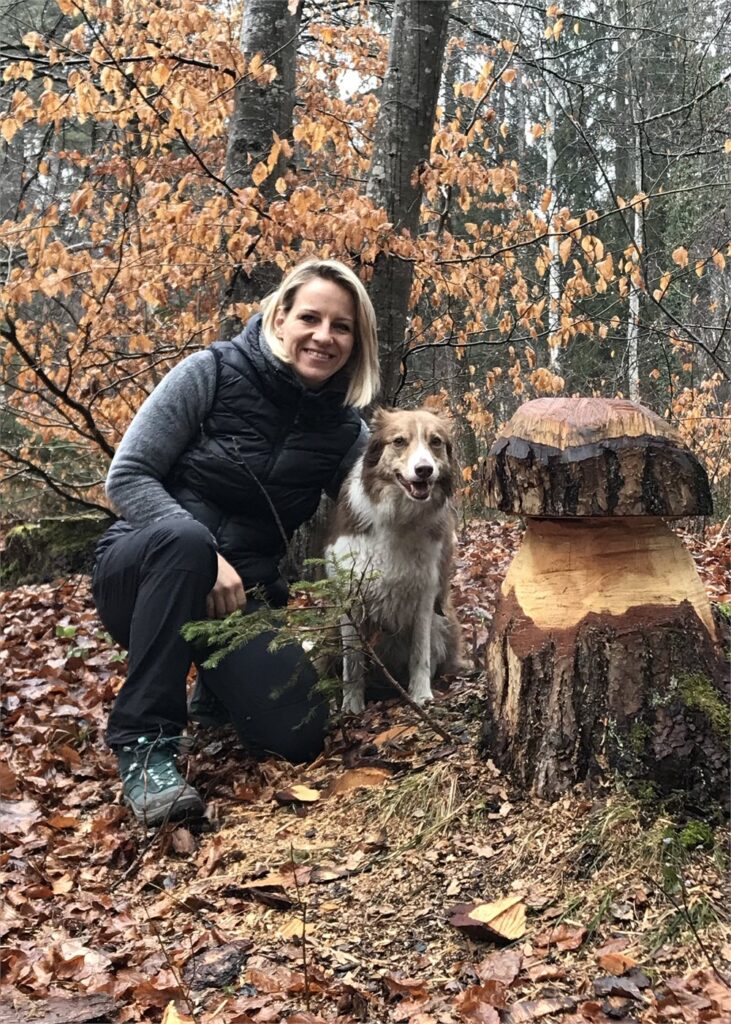 Alexandra Ritter hockt im herbstlichen Laubwald neben ihrem Hund und einer aus einem Baumstrunk geschnitzten Pilzfigur