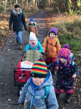 Eine Betreuerin und Kinder ziehen einen Leiterwagen über den Waldweg