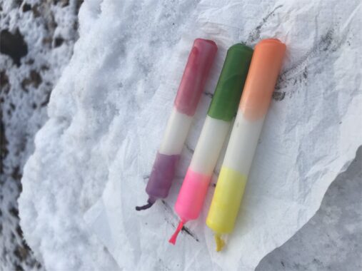 Drei selbst gegossene Kerzen in Rot, Grün und Orange liegen im Schnee