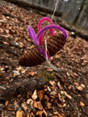 Ein Tannzapfen mit pinken Pfeifenputzern hängt an einer Schnur